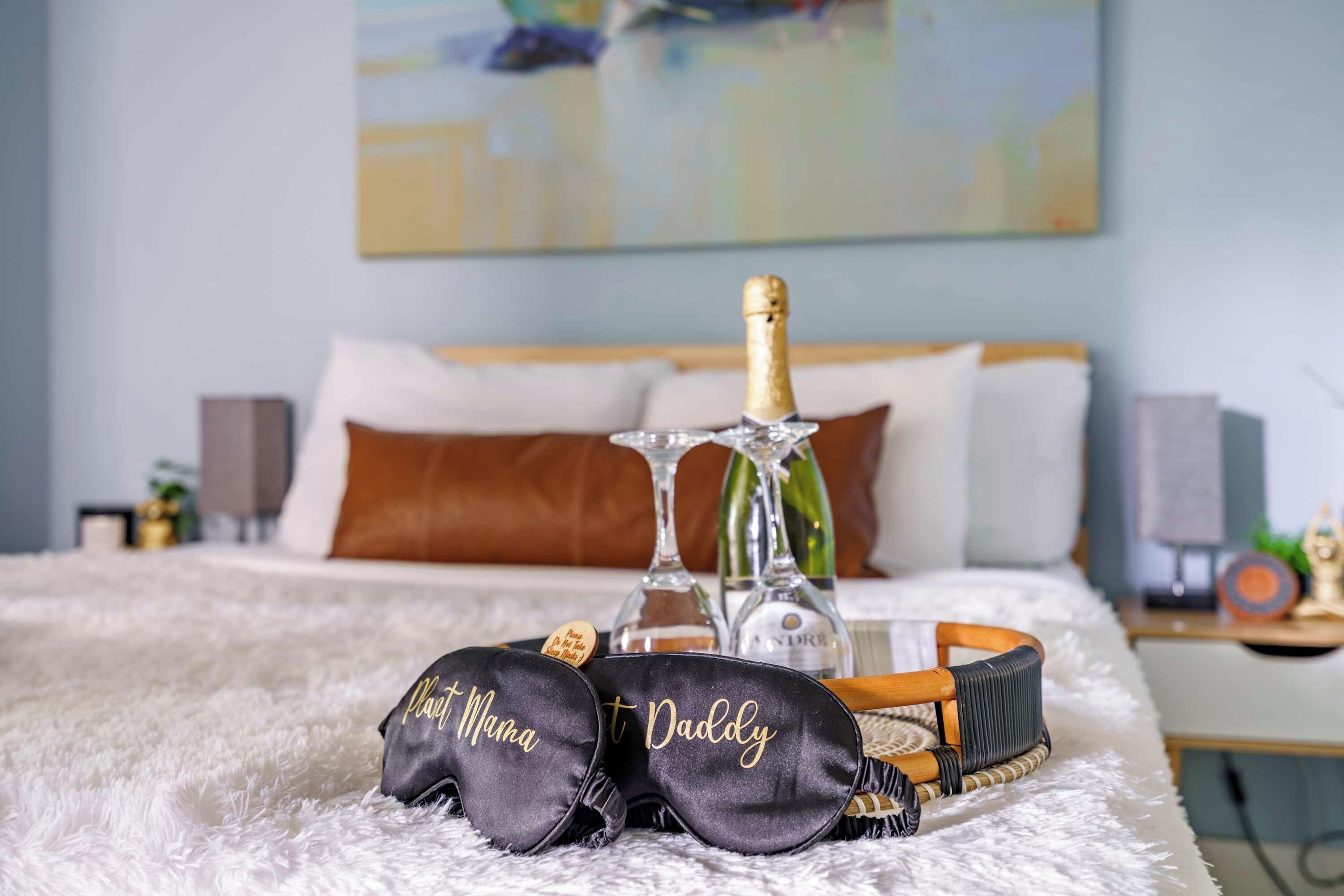 Ein Bett mit einer flauschigen weißen Decke, eine Champagnerflasche, zwei Gläser und ein Korbwesen mit schwarzen Kissen, auf denen 'Papa' und 'Mama' stehen.