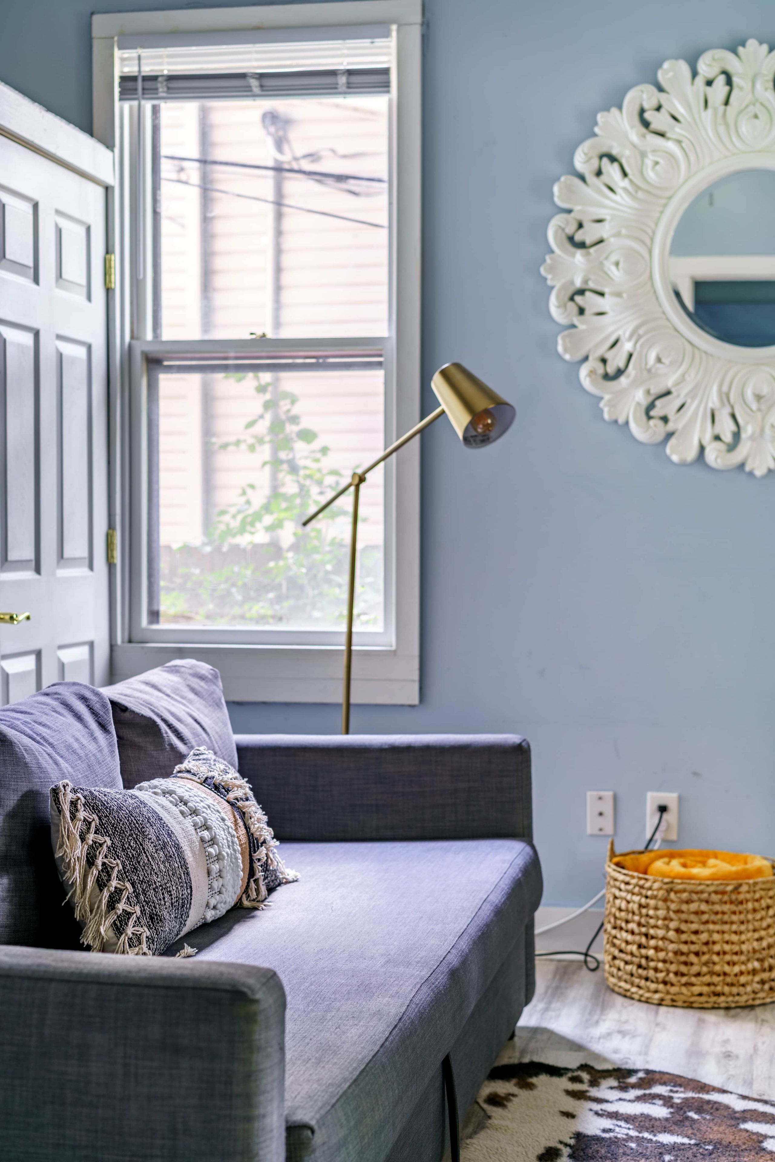 Een kamer met een grijze bank, een decoratieve spiegel en een lamp.