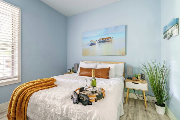 A bedroom with a bed, a painting, a nightstand, a potted plant, and a window with blinds.