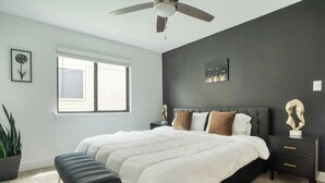 Une chambre à coucher avec un lit, des tables de chevet, un ventilateur de plafond, une fenêtre et une plante en pot.