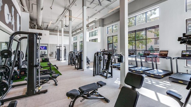 Un gimnasio amplio con diversos equipos de ejercicio, ventanas grandes y un ventilador de techo.