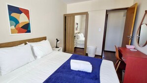 A hotel room with a bed, a blue blanket, a wooden headboard, a white pillow, a nightstand, a lamp, a mirror, a red desk, a white basket, and a colorful abstract painting on the wall. - CONAZ0100 - Edifício Larissa (Salvador)