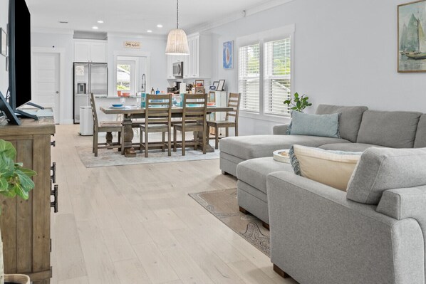 A modern kitchen with white cabinets, a dining table with chairs, and a grey sofa in the living area.