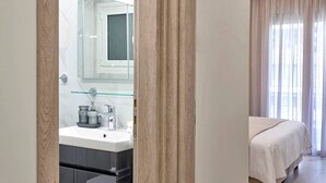 A modern hotel room with a bathroom featuring a glass shower enclosure, a white sink, and a mirror. The room has a bed with white bedding and a large window with curtains.