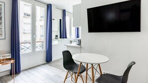 A modern room with a large window, a flat-screen TV, a round table with two chairs, and a small kitchenette area.