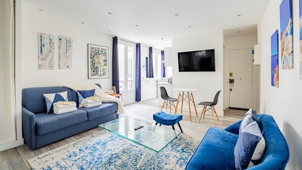 A modern living room with a blue sofa, a glass coffee table, and a blue armchair. There is a TV mounted on the wall and a small dining area with two chairs.