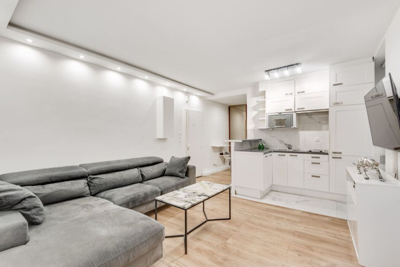 Ein modernes Wohnzimmer mit einem grauen Ecksofa und einem kleinen Couchtisch, eine Küche mit weißen Schränken und einem Einbauofen, sowie ein Flachbildfernseher an der Wand.