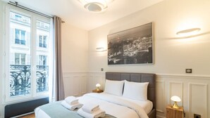 Habitación de hotel con cama, dos lámparas de noche, ventana con vista a edificios y un cuadro de paisaje urbano en la pared.