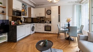 Una cocina moderna con armarios blancos, horno empotrado y microondas. Cuenta con una zona de comedor con mesa y sillas, y un sofá.