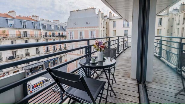 Un balcon avec une table et des chaises, un vase de fleurs et une vue sur les bâtiments environnants.