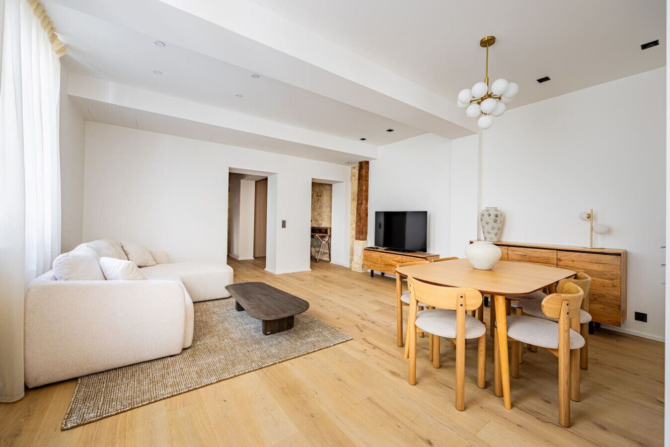 Un salon moderne comprenant un canapé, une table basse, une table à manger et des chaises. On y trouve un téléviseur posé sur un meuble en bois, et un lustre est suspendu au plafond.