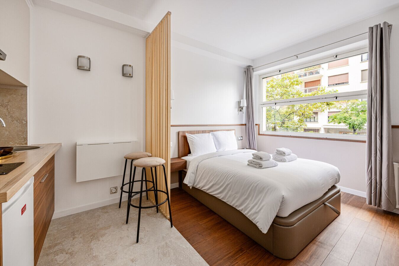 Een moderne hotelkamer met een bed, een kleine kitchenette, een houten scheidingswand en een raam met uitzicht op gebouwen en bomen.