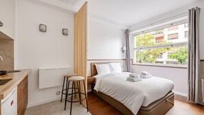 A modern hotel room with a bed, a small kitchenette, a wooden divider, and a window with a view of buildings and trees.