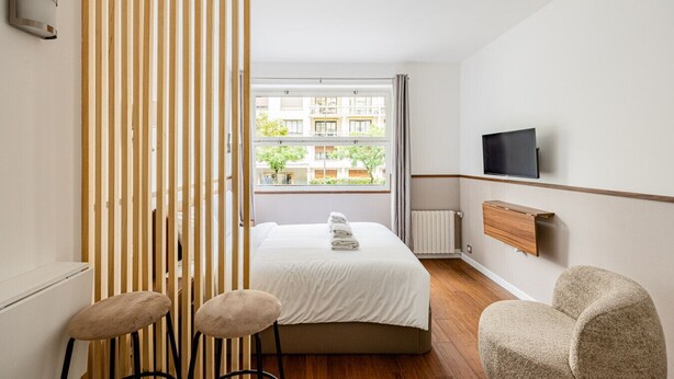 A hotel room with a bed, a TV, a wooden partition, and a view of buildings outside.