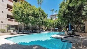 A swimming pool with lounge chairs and umbrellas, surrounded by trees and a building with balconies.