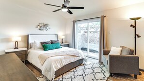 Un dormitorio con cama, mesitas de noche, una silla, ventilador de techo y un ventanal grande.