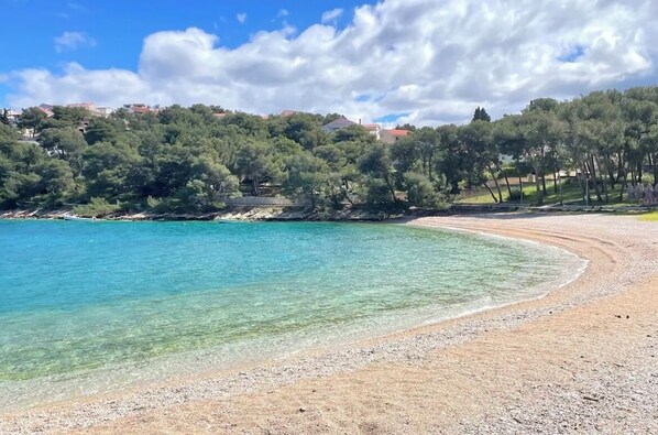 A beach with clear turquoise water, a pebbly shore, and a backdrop of trees and buildings.