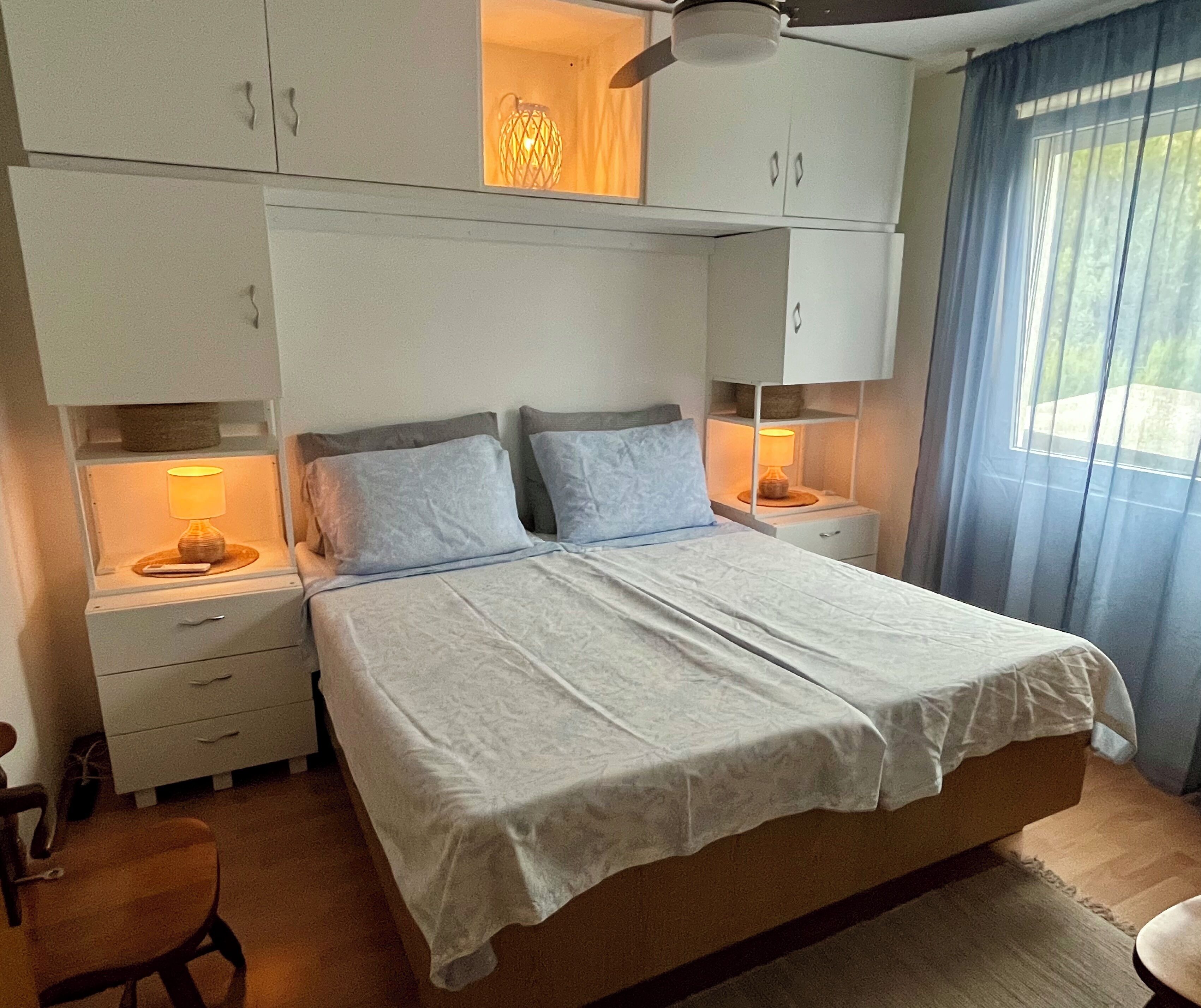 A bedroom with a large bed, two bedside tables with lamps, a ceiling fan, and a window with curtains.