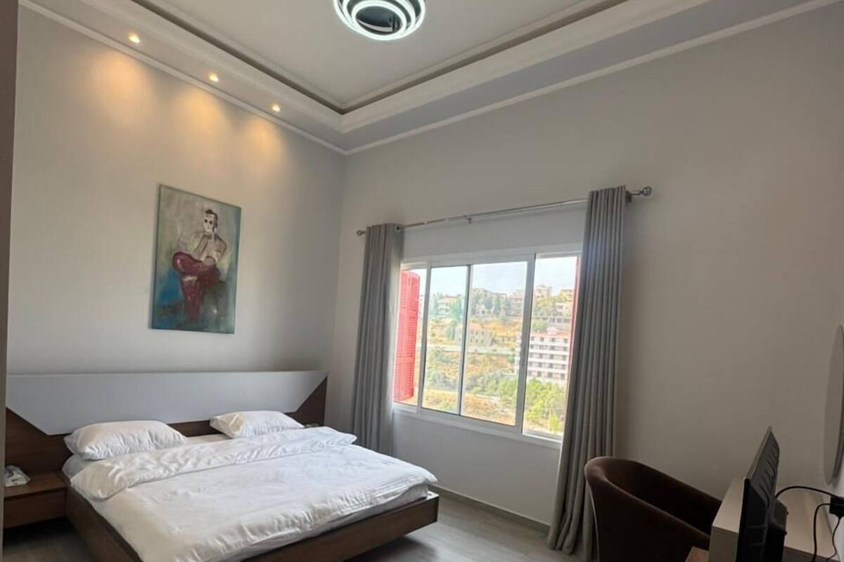 A modern hotel room with a large bed, a desk with a computer, a chair, and a painting on the wall.