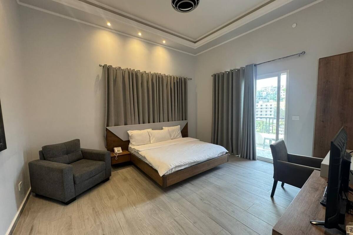 A modern hotel room with a large bed, a desk, a chair, and a view of the city through the window.