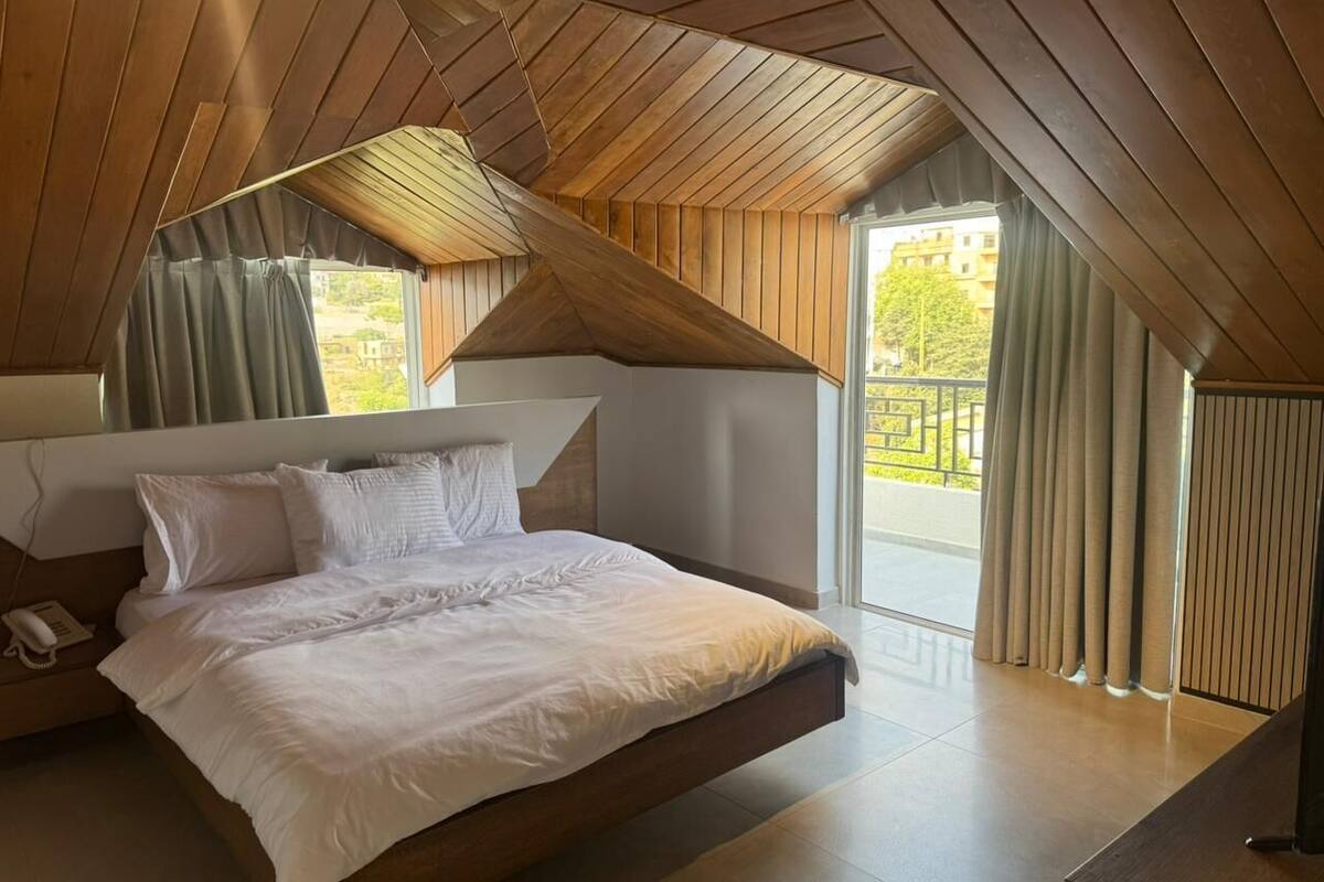 A bedroom with a sloped ceiling, a large bed, a bedside telephone, and a view of the outdoors through a window with curtains.
