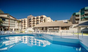 Una piscina con cubierta de baldosas, un área de estar techada y un edificio con varios balcones al fondo.