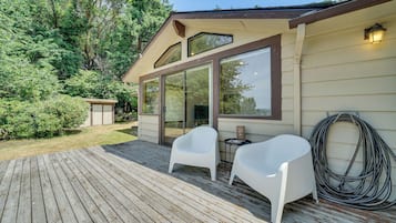 A wooden deck with two white chairs and a table, a covered patio area, and a garden with trees and shrubs.
