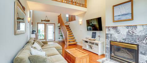 A spacious living room with a high ceiling, a wooden staircase, a fireplace, and a large window.