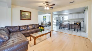 A living room with a brown leather sofa, a glass coffee table with flowers, and a ceiling fan. A kitchen is visible in the background with white cabinets and a bar area. - Blue Ridge Retreat on 10-Acre Farm: Rural Reset! (Blue Ridge)