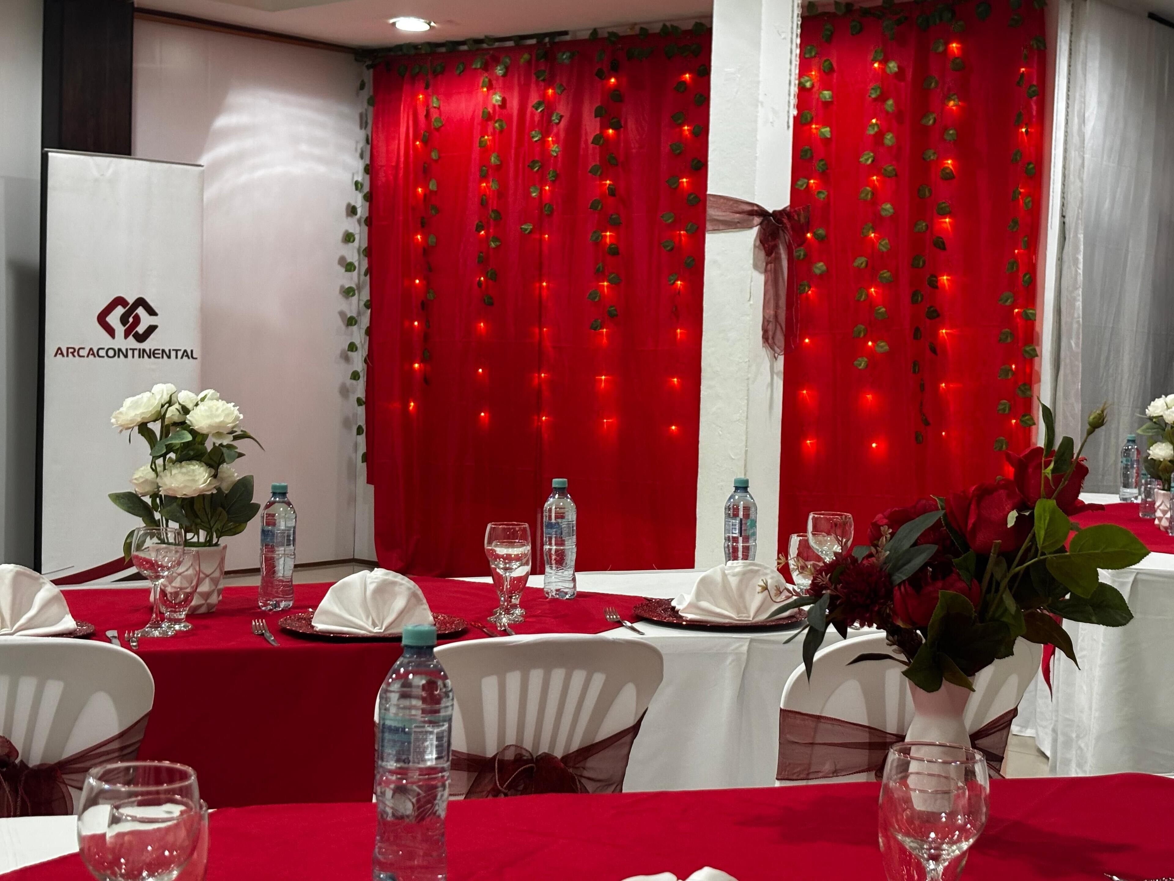 A banquet hall with tables set for a formal event, featuring red curtains with leaf decorations and string lights, white tablecloths, and floral centerpieces.