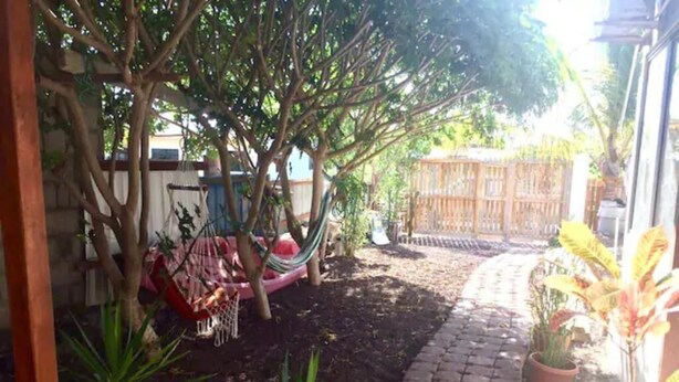 Uma área de jardim com caminho pavimentado, plantas em vasos, uma rede e cerca de madeira.