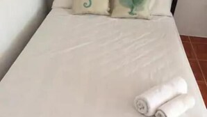 Uma cama com colchão branco e duas almofadas com estampas verdes, uma toalha branca enrolada sobre a cama e uma estrutura de metal.