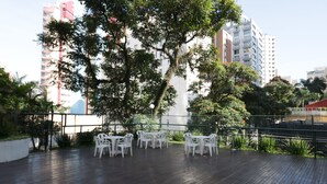 An outdoor patio with white plastic chairs and tables, surrounded by greenery and a metal railing.