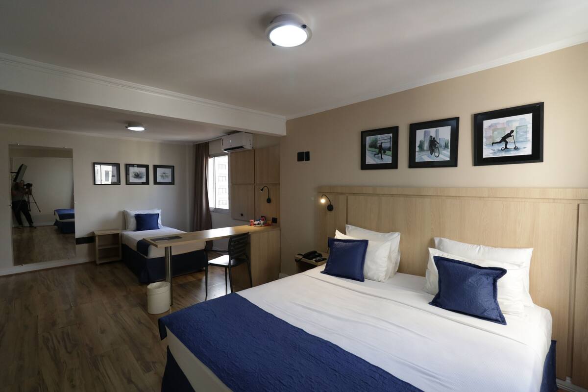 Une chambre d’hôtel avec un grand lit, un bureau et un miroir. Les murs sont ornés de tableaux encadrés et le plafond est éclairé par un luminaire.