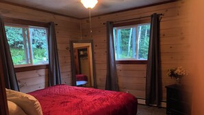 A bedroom with a red bedspread, wooden walls, and two windows with curtains. - Soothing cabin at Great sacandaga lake come enjoy the peace in a rural area. (Hadley)
