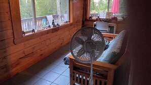 A wooden cabin interior with a fan, a sofa, and a window with a view of a deck and trees. - Soothing cabin at Great sacandaga lake come enjoy the peace in a rural area. (Hadley)