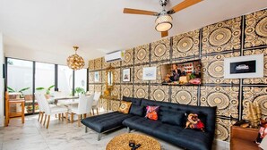 A modern living room with a black sectional sofa, a round wicker coffee table, and a dining area with a white table and chairs. - Charming & Comfortable House, 15 min to Downtown (Playa del Carmen)