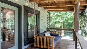 Terraza de madera con una mecedora y un sofá, ventanas amplias y vistas a árboles.