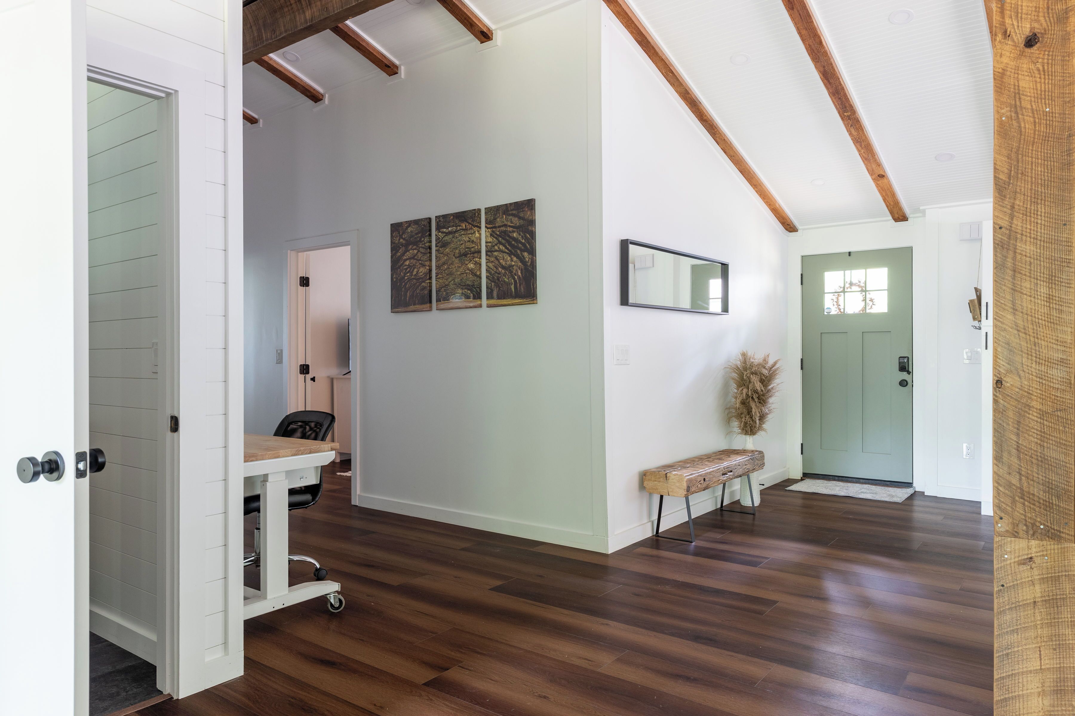 A spacious room with hardwood flooring, a white door, a bench, and a wall-mounted mirror.