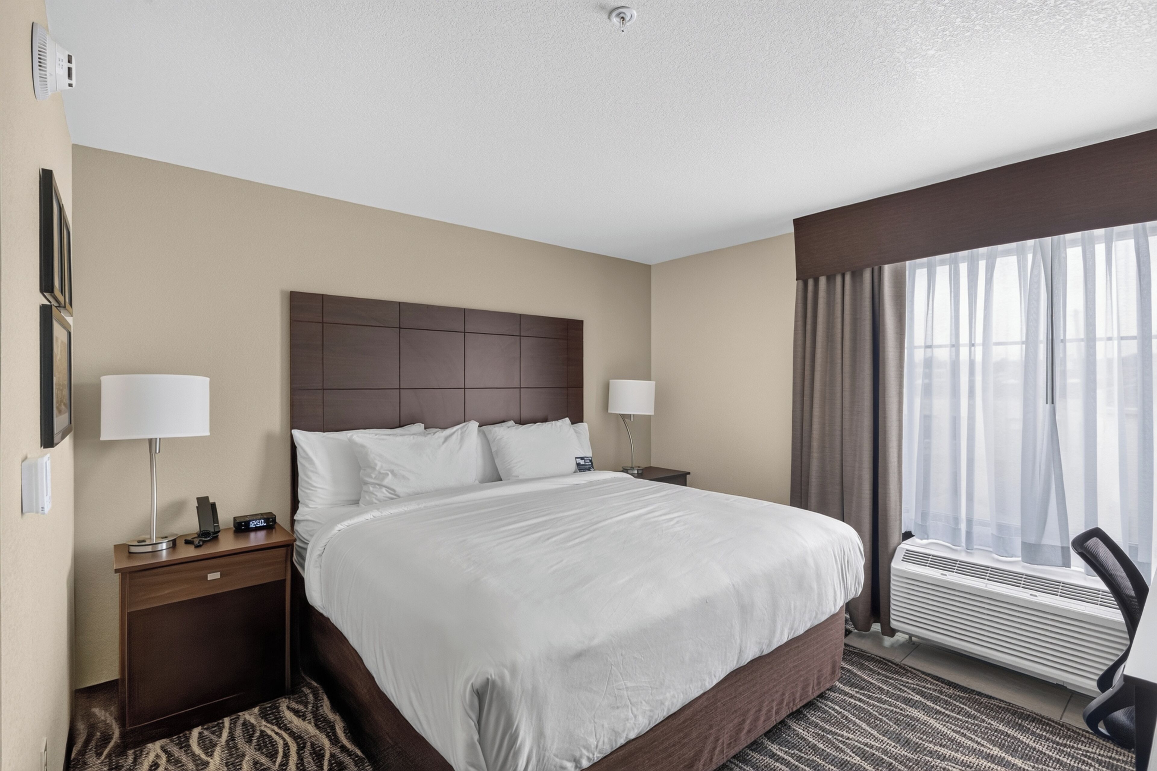 A hotel room with a large bed, two bedside tables, a nightstand, a wall-mounted air conditioner, and a window with curtains.