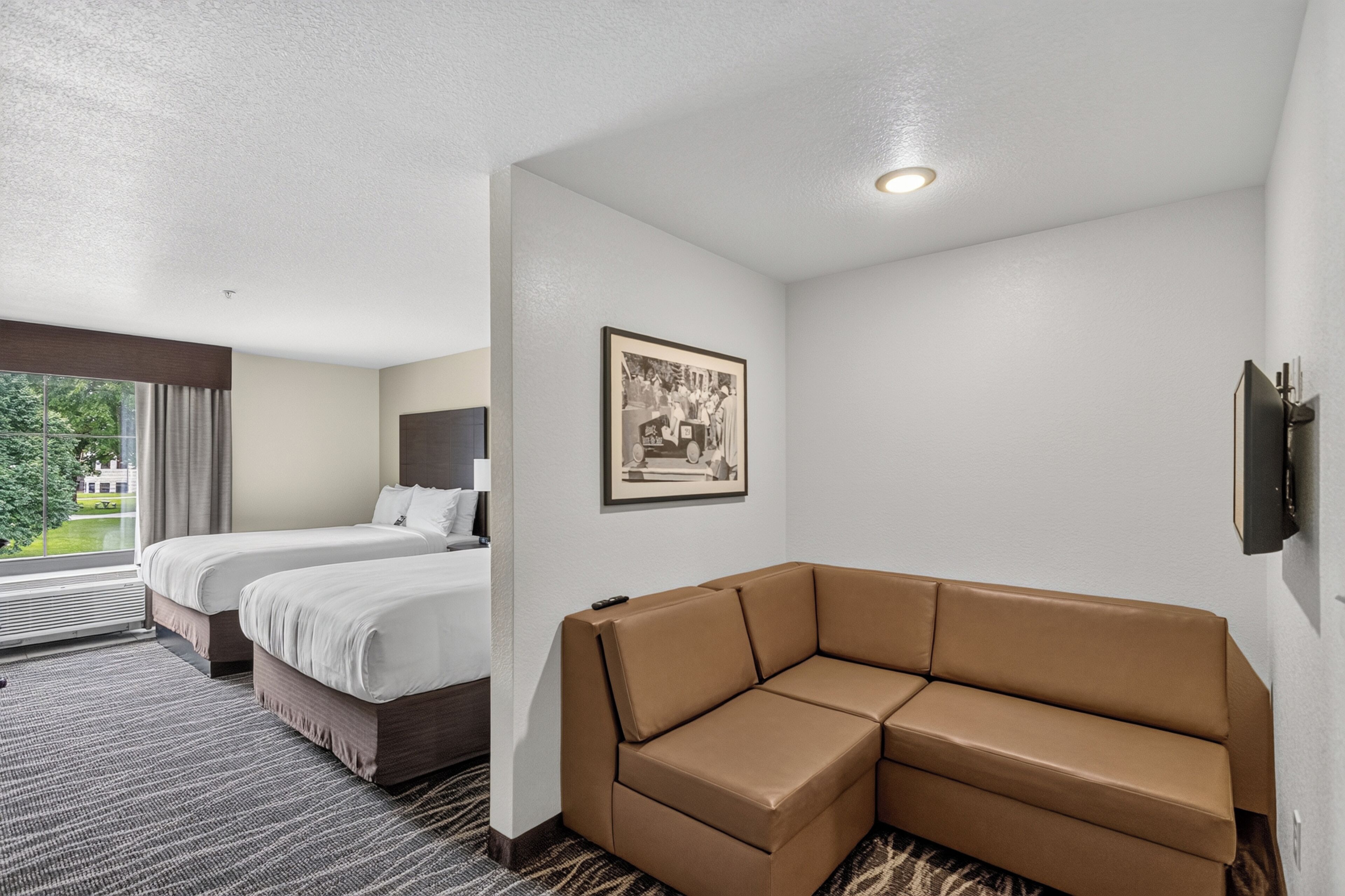 A hotel room with two beds, a brown sofa, a framed picture on the wall, and a window with curtains.