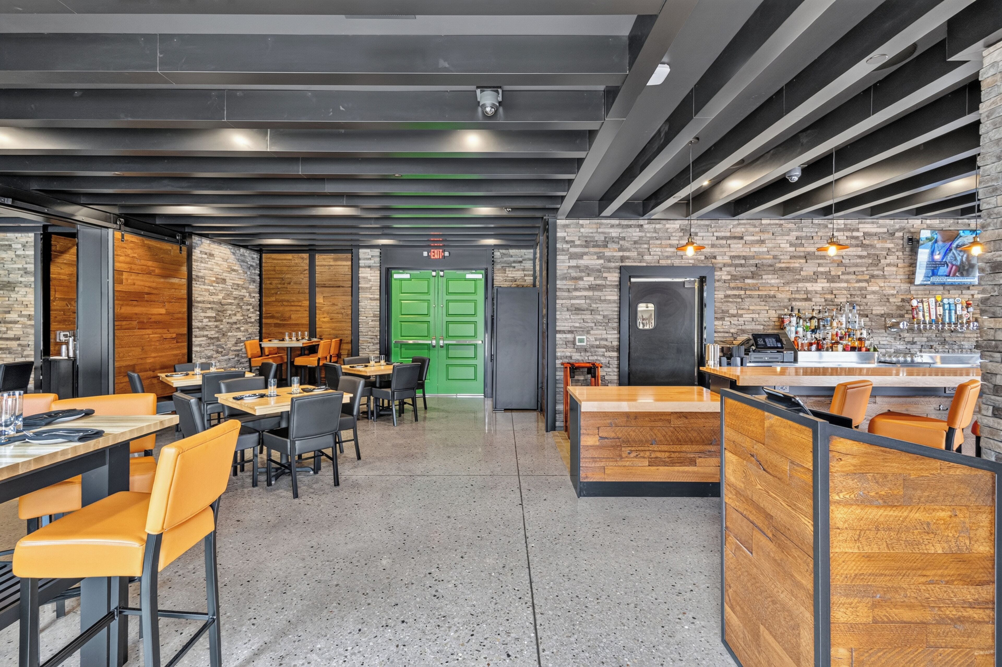 A modern restaurant interior with high tables, chairs, a bar area, and a green exit door.