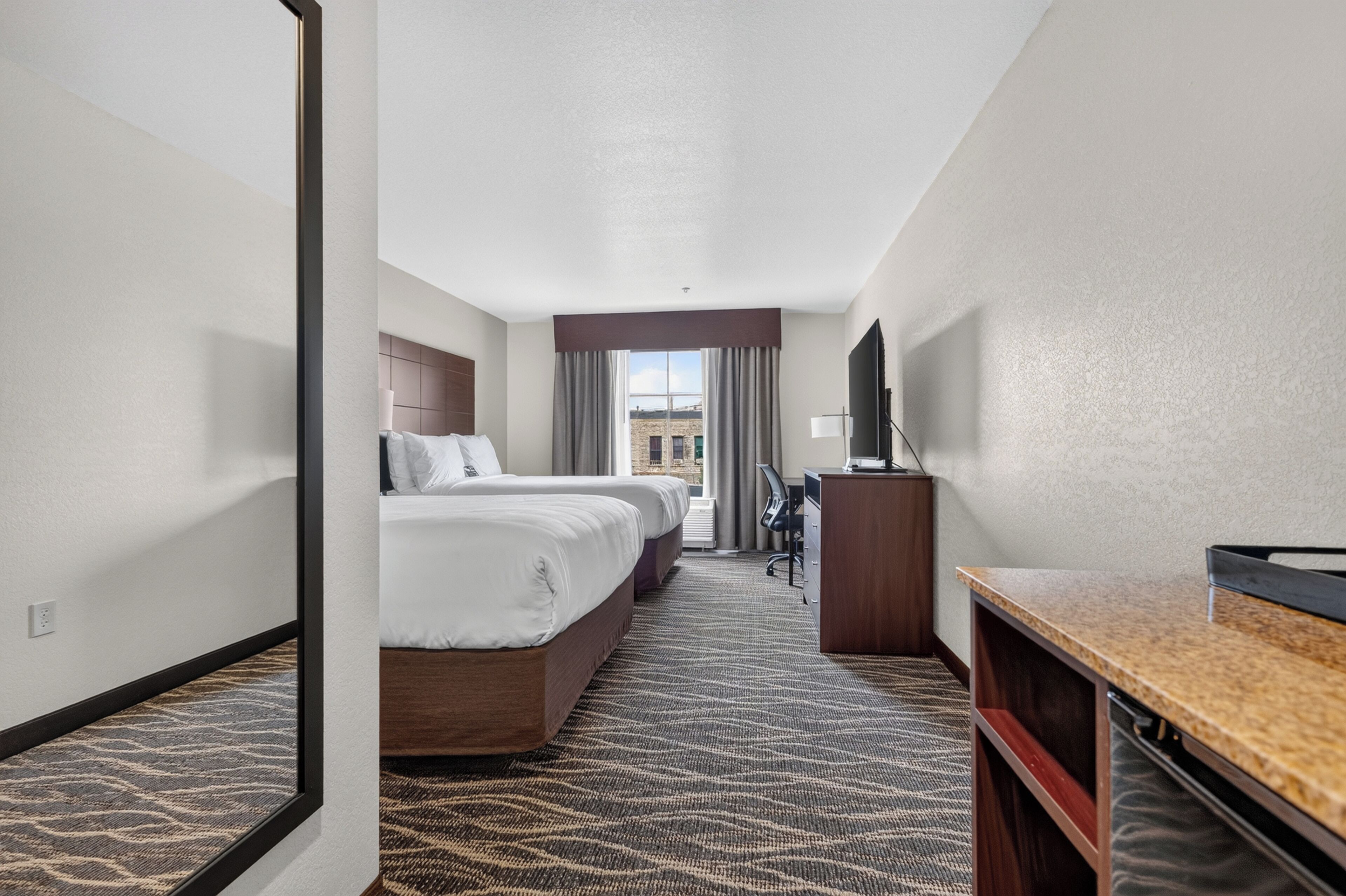 A hotel room with two beds, a desk, a TV, and a view of the outside.