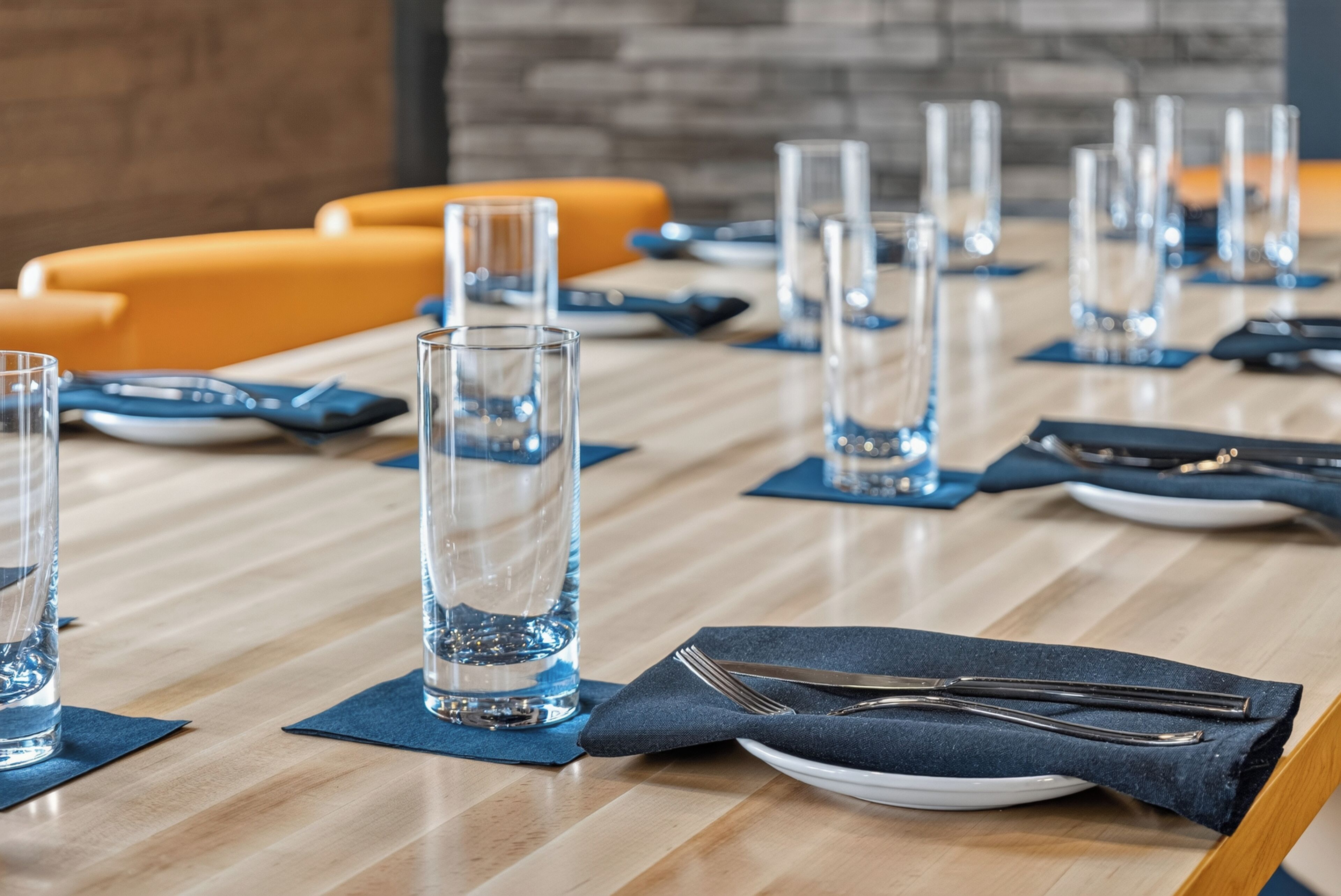 Una mesa de conferencias dispuesta con vasos transparentes, platos y cubiertos, con sillas anaranjadas al fondo.