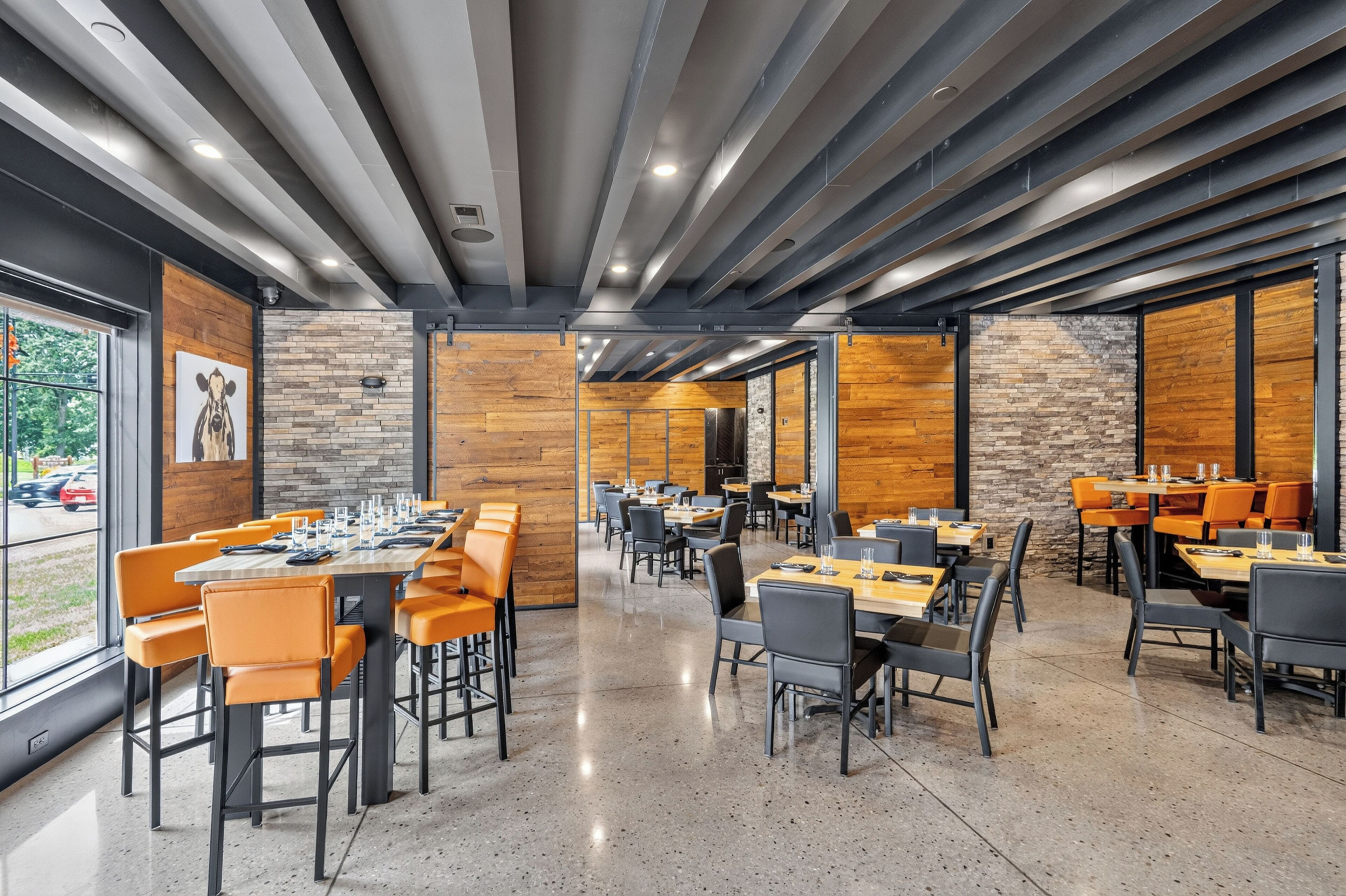 Un interno ristorante moderno con soffitti alti, travi a vista e un mix di pannelli murali in mattoni e legno. Tavoli disposti per la ristorazione con sedie.