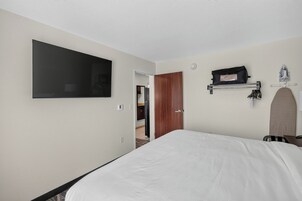 Habitación de hotel con una cama, un televisor montado, un perchero y una puerta que da a un baño.