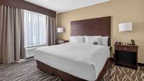 A hotel room with a large bed, a bedside table, a lamp, a clock, and a window with curtains. - Cobblestone Hotel & Suites - Waukesha/West Milwaukee (Waukesha)