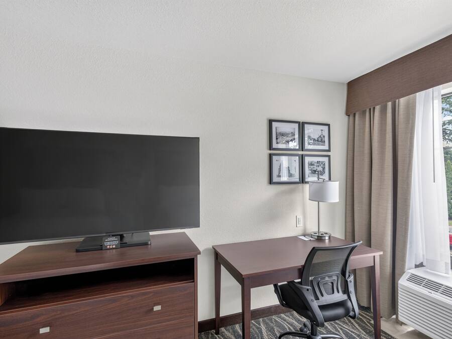 A hotel room with a flat-screen TV, a desk, a chair, a lamp, and a window with curtains.
