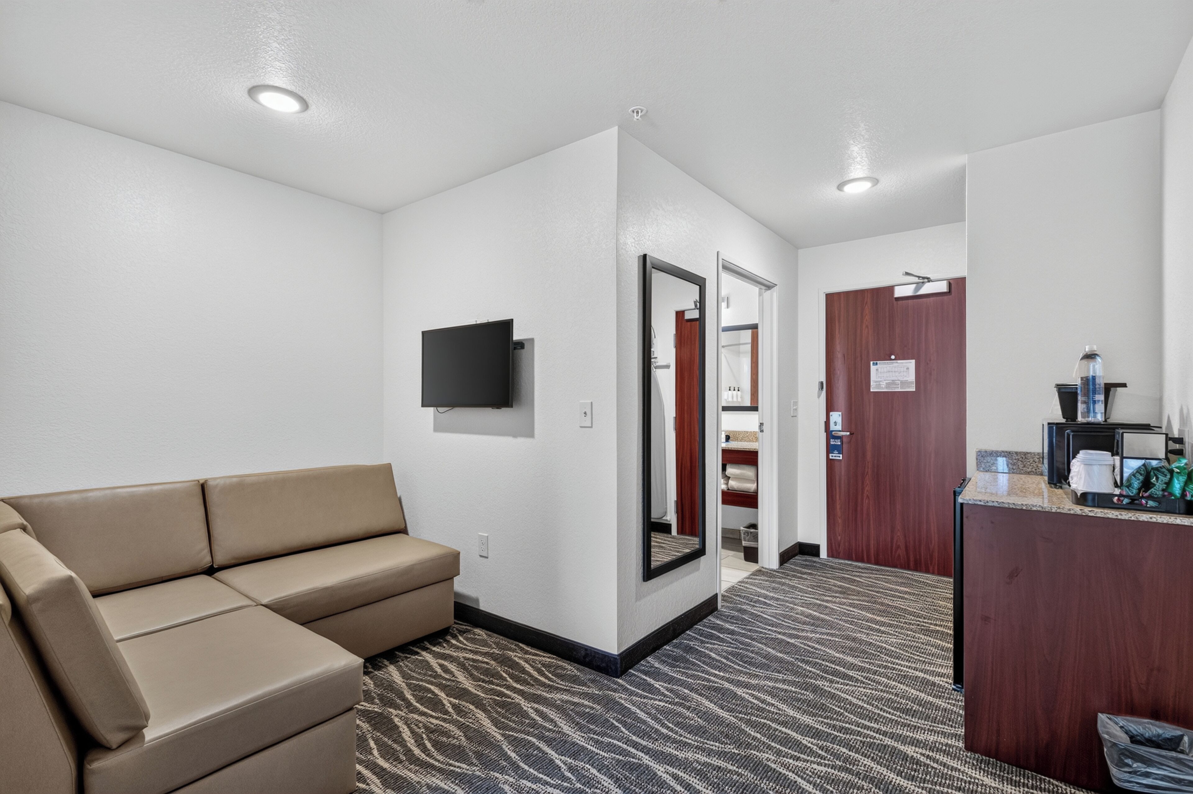 Habitación de hotel con sofá, televisor de pantalla plana, espejo, puerta que da a un baño y mostrador de recepción con servicios.