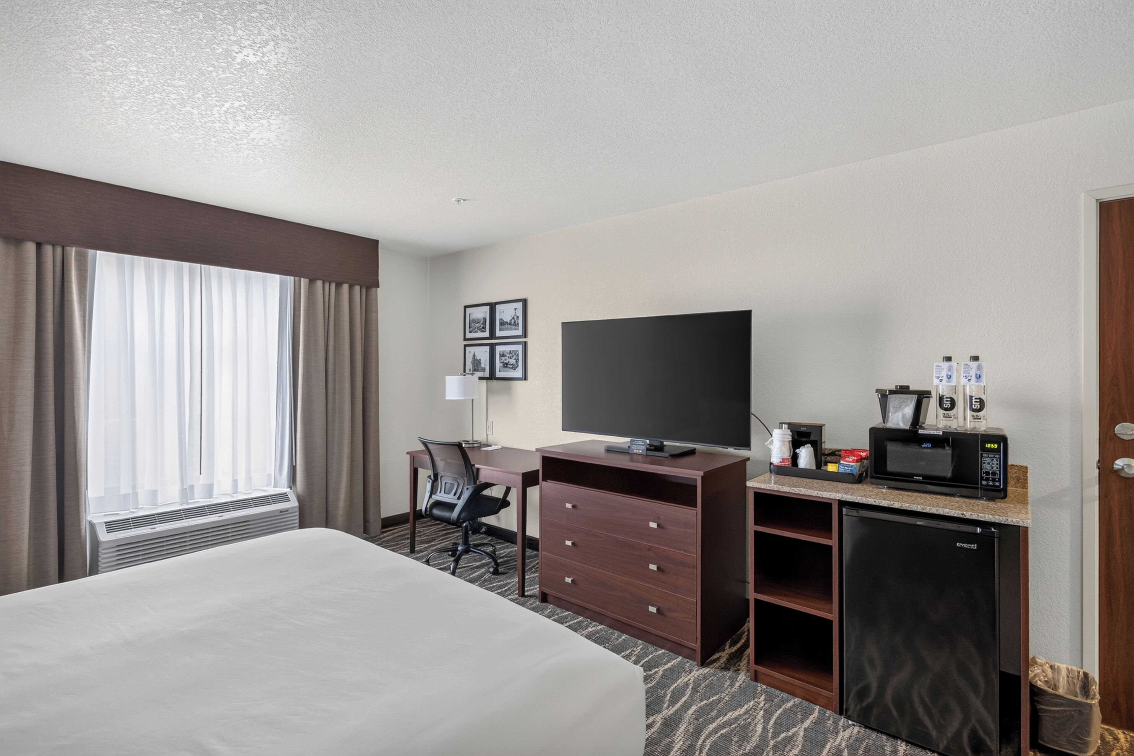 A hotel room with a bed, desk, chair, television, minibar, and a window with curtains.